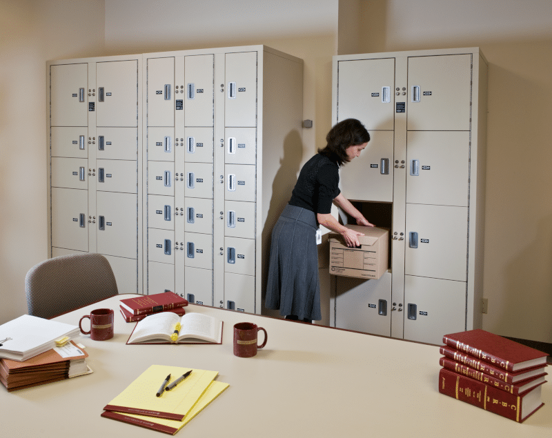 Pass-back evidence lockers. Woman is putting item in locker behind a table covered in books and paperwork